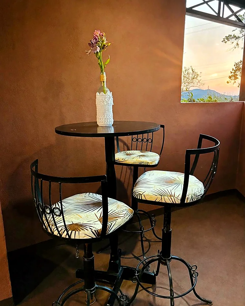 Mesa alta com três bancos junto à parede terracota, com o morro ao fundo pela janela
